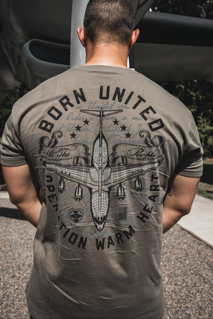 A man stands outdoors with his back to the camera, wearing the Born United Private Label Operation Warm Heart–Olive Limited Edition Tee featuring a military cargo plane graphic and text supporting our heroes.