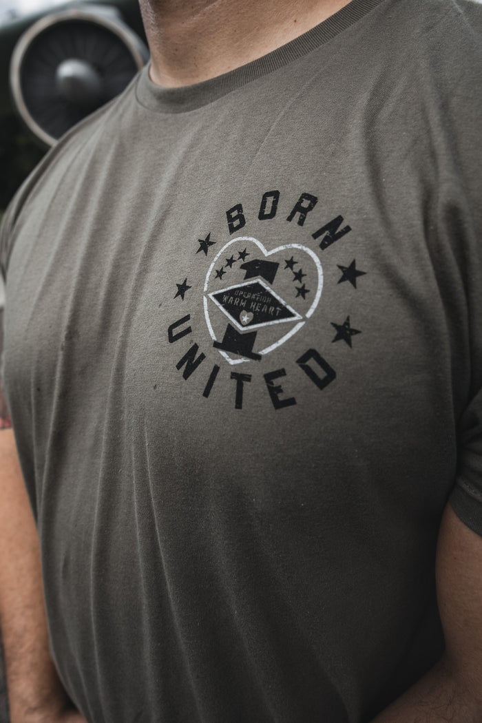 A person wears the Born United Private Label Operation Warm Heart-Olive Tee, featuring a circular BORN UNITED logo with a star and banners reading HANDCRAFTED and WARE HEART. A blurred engine in the background shows support for our heroes.