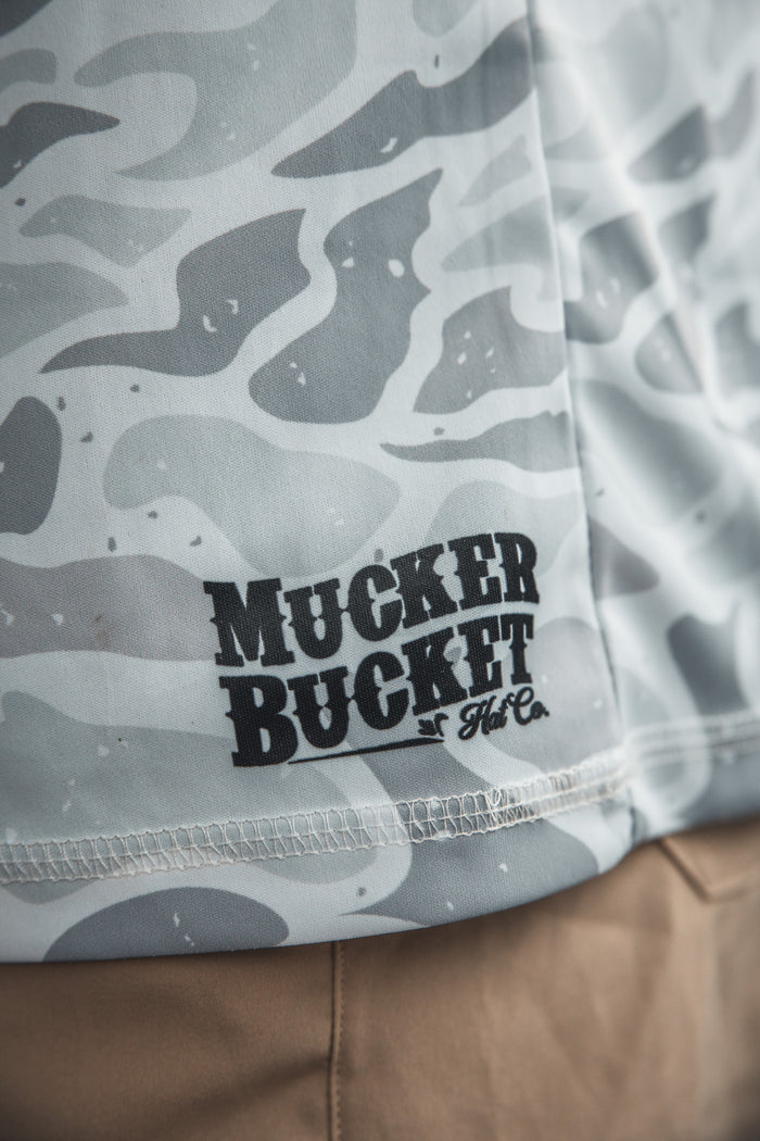 Close-up of the Mucker Bucket Performance Hoodie in Ghost Camo, featuring a light gray and white camouflage pattern with the brand printed in black, draped over tan pants.