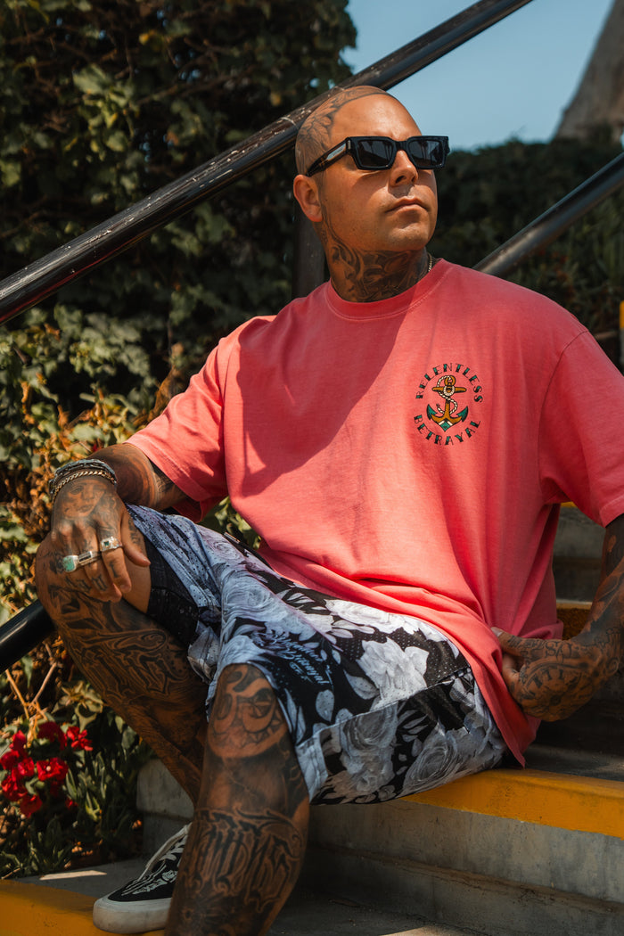 A tattooed man in sunglasses, a pink T-shirt, and Relentless Betrayals Premium Mesh Shorts | 5- Beauty in Darkness sits on outdoor stairs amidst greenery, sunlight creating dramatic shadows around him.