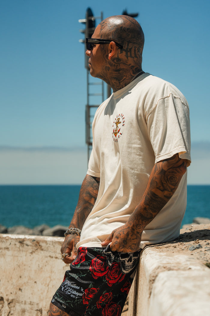 A tattooed man in sunglasses and a white T-shirt wears Relentless Betrayal’s Premium Mesh Shorts | 5- Thorn Amongst Roses as he leans on a concrete wall by the sea under a clear blue sky.