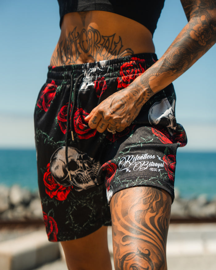 A tattooed person stands by the ocean on a sunny day, wearing Relentless Betrayals Premium Mesh Shorts | 5- Thorn Amongst Roses with red rose and skull designs, paired with a black crop top and hand resting on the waistband.