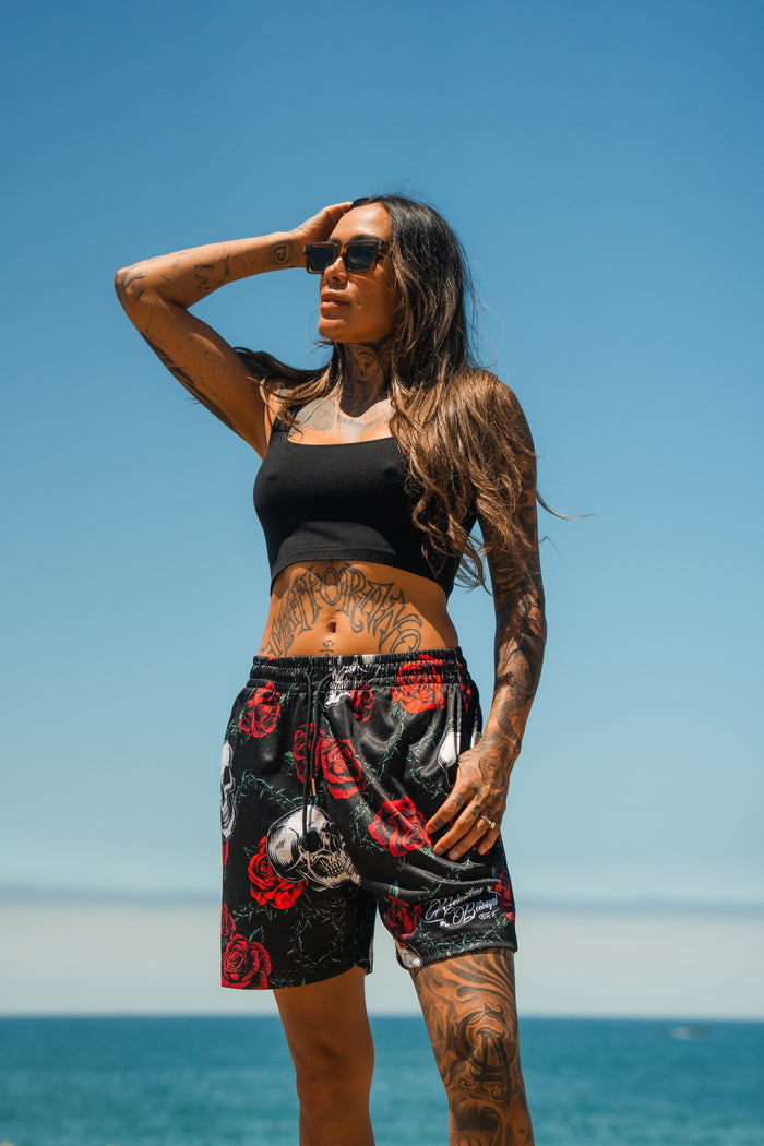 A woman with long dark hair and tattoos poses by the ocean under a clear blue sky, wearing sunglasses, a black crop top, and Relentless Betrayal Premium Mesh Shorts | 5- Thorn Amongst Roses.
