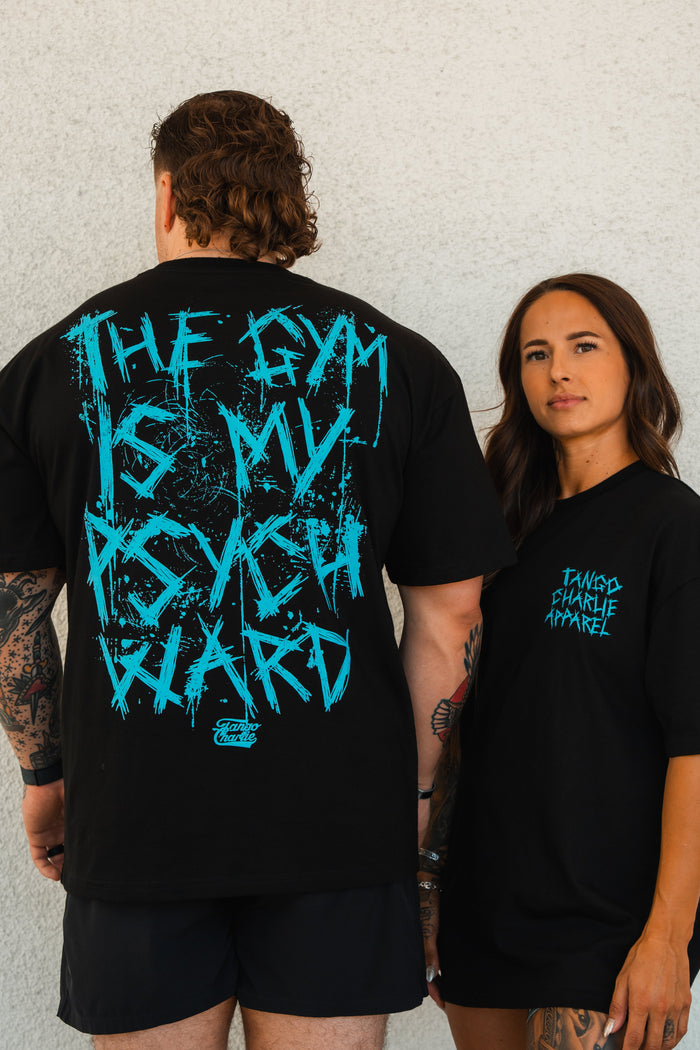 A man and a woman wear Tango Charlie Apparels Psych Ward - Premium Oversized Tee in black; his says THE GYM IS MY PSYCH WARD in bold blue, while hers features the brand logo in smaller blue text on heavyweight cotton.