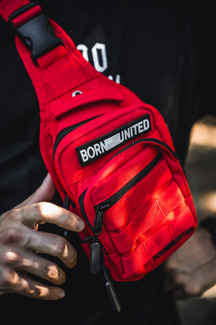 A person wearing the Red Reaper Tactical Sling-Pac (4L) by Born United Private Label, featuring a bold black-and-white "BORN UNITED" patch, stands against a dark shirt and holds the compact red bag with their left hand.