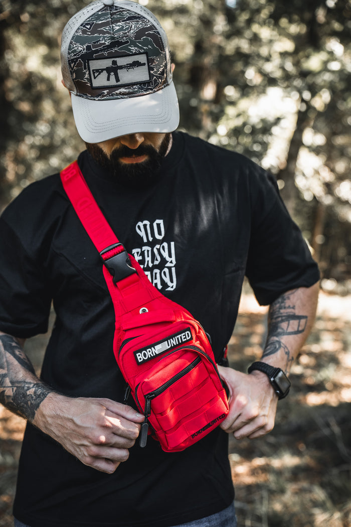 A bearded man in a camo cap and black T-shirt adjusts the Born United Private Label Red Reaper Tactical Sling-Pac - 4L outdoors, with trees and sunlight blurred in the background.