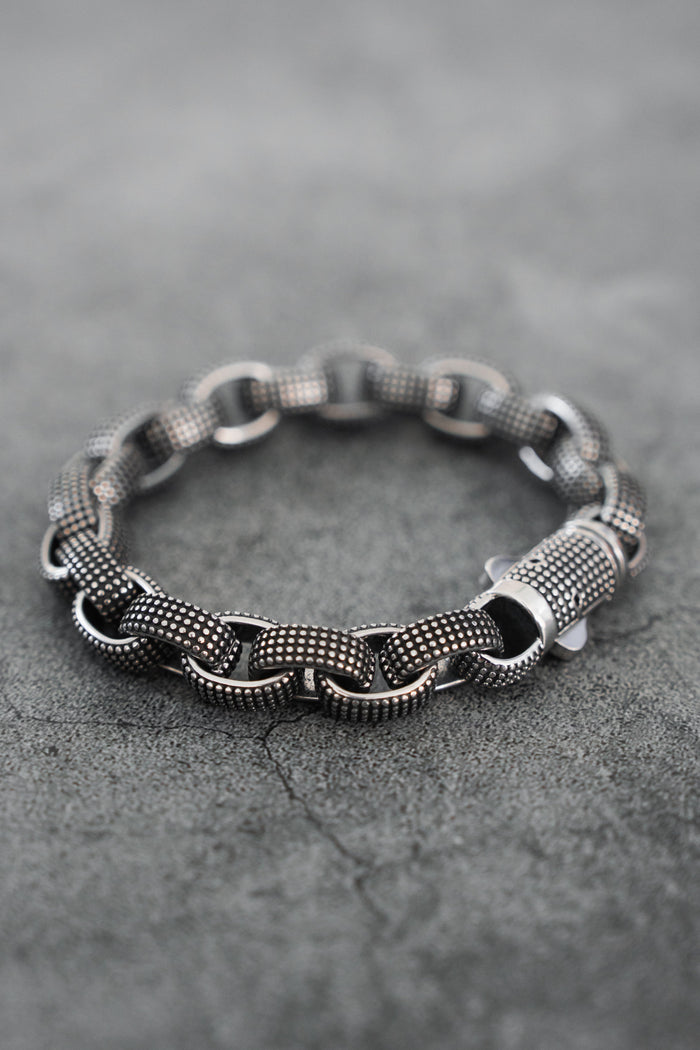 The Relentless Betrayal Seeker Bracelet, hand-forged from stainless steel with large interlocking links and a textured silver-tone finish, is shown on a gray concrete surface.