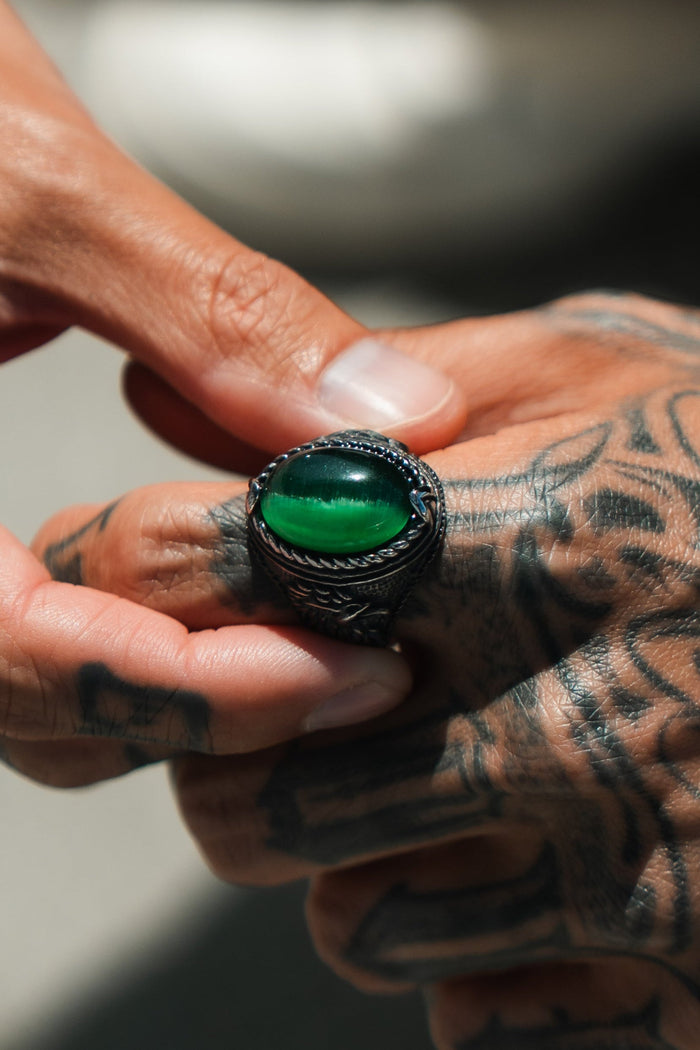 Tattooed hands are shown as one places a Relentless Betrayals Serpents Eye Ring with a large green gem onto the finger, set against an artistically blurred stainless steel background.