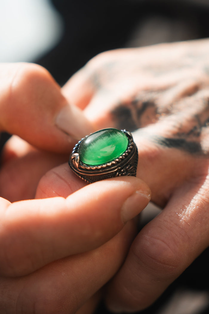 The close-up shows a tattooed hand gently holding Relentless Betrayals Serpents Eye Ring, featuring an ornate dark metal band and a large oval green gemstone.