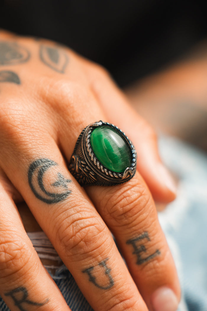 A close-up reveals a tattooed hand adorned with a striking Relentless Betrayal Serpents Eye Ring, featuring a polished green stone. The stainless steel band fits comfortably, highlighting intricate finger tattoos and adding an air of mystique as the hand rests on a surface.