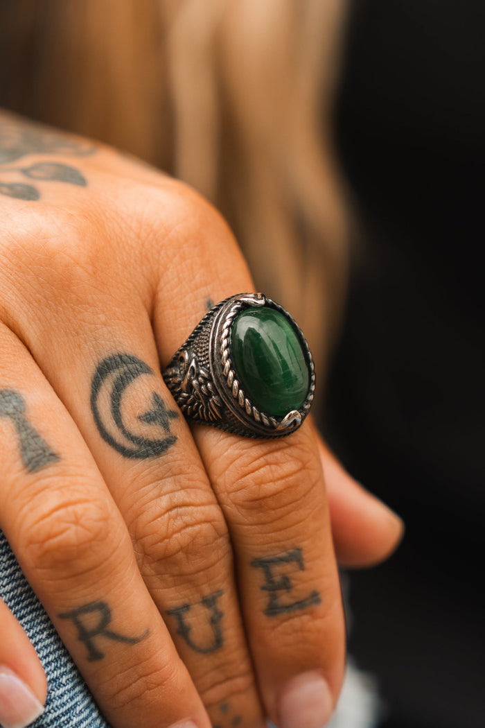 A tattooed hand flaunts the Serpents Eye Ring from Relentless Betrayal, boasting a polished green stone set in an intricate metal band. The lettered tattoos enhance its mystique, while a blurred background accentuates its allure.
