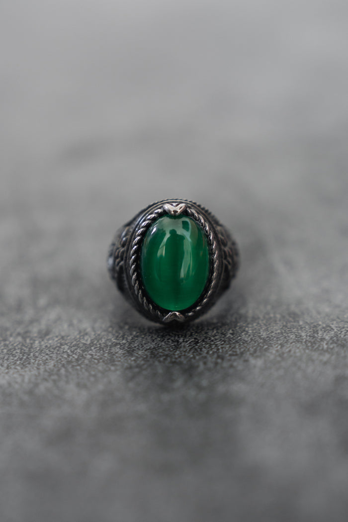 The Serpents Eye Ring from Relentless Betrayal, featuring a large oval green gemstone at its heart and an ornate silver rope-patterned band, rests elegantly on a smooth gray surface.