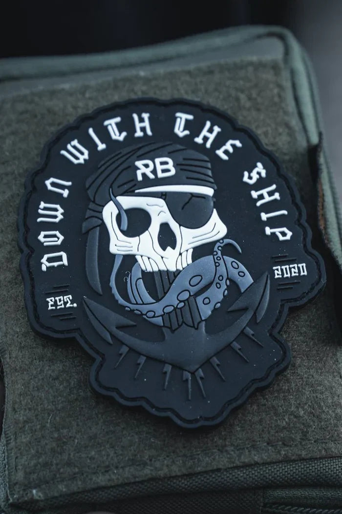 The Relentless Betrayal Ship Wreck PVC Patch features a skull in a bandana labeled RB, biting a chain, and DOWN WITH THE SHIP in bold white on black, symbolizing survival, attached to green fabric.