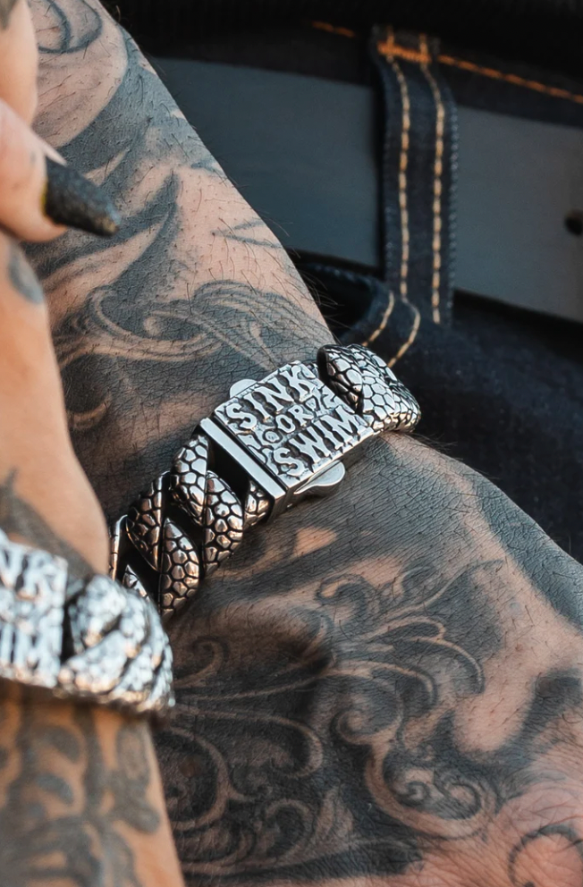 A tattooed arm showcases the Relentless Betrayal Sink or Swim Bracelet, featuring a textured silver chain and an engraved plate reading Sink or Swim. The wearers dark clothing with visible belt loops enhances this bold statement piece.