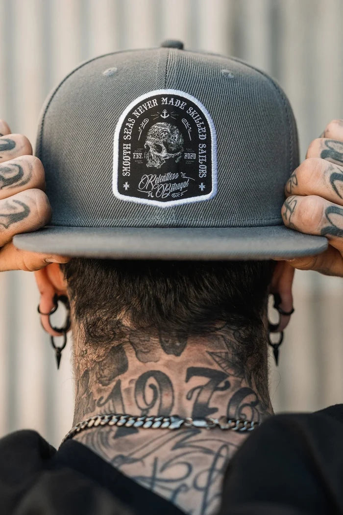 A tattooed person is seen from the back, holding the Skilled Sailor Snapback by Relentless Betrayal with both hands. The gray polyester cotton blend cap features embroidered nautical patches and text.
