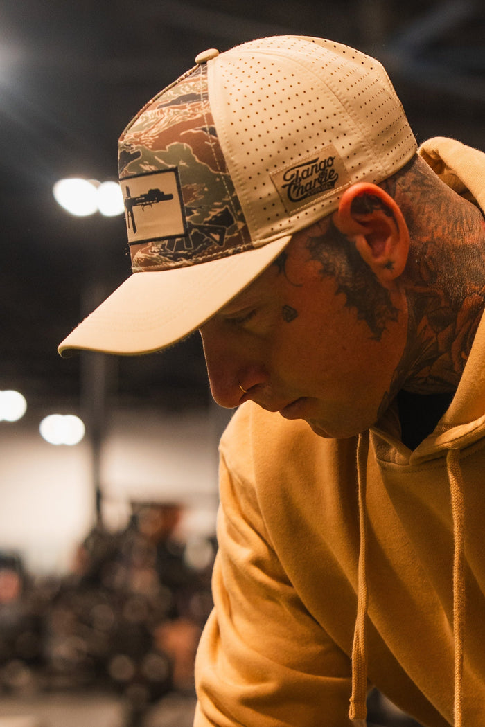 A man with face and neck tattoos looks down in soft indoor light, wearing a tan hoodie and the Snipes & Stripes Hydro Snapback Hat by Tango Charlie Apparel. The blurred background features lights and indistinct shapes.