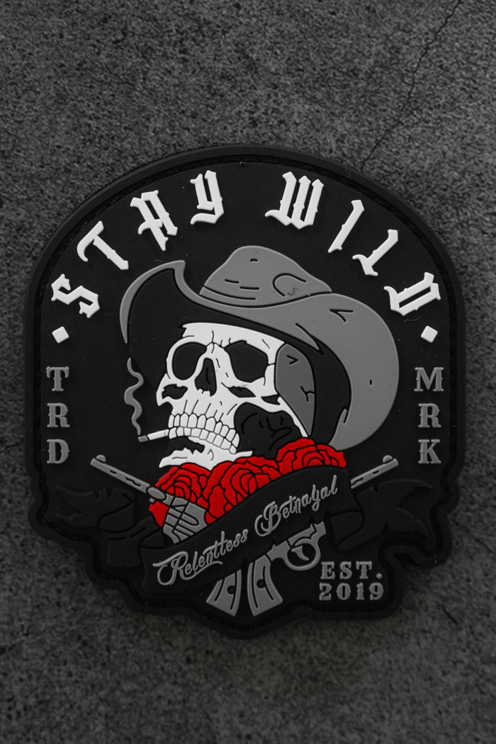 The Stay Wild Patch by Relentless Betrayal is a black Velcro patch featuring a skull in a cowboy hat smoking a cigarette, red roses, crossed revolvers, and the text “STAY WILD,” “Relentless Betrayal,” and “EST 2019.”.