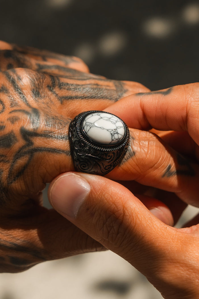 Tattooed hands adjust the Relentless Betrayal Storm Cloud Ring, crafted from blackened metal and set with a large white stone veined in gray. Details of skin and ring are highlighted in natural light.