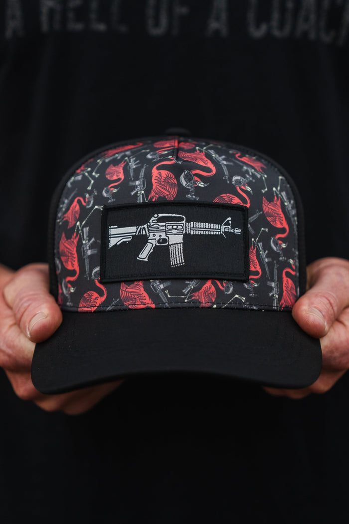 An individual holds a Tango Charlie Apparel Tactimingo Hydro snapback hat, featuring a sweat-resistant red and gray design with guns and buffalo, centered by a patch illustrating a white rifle.