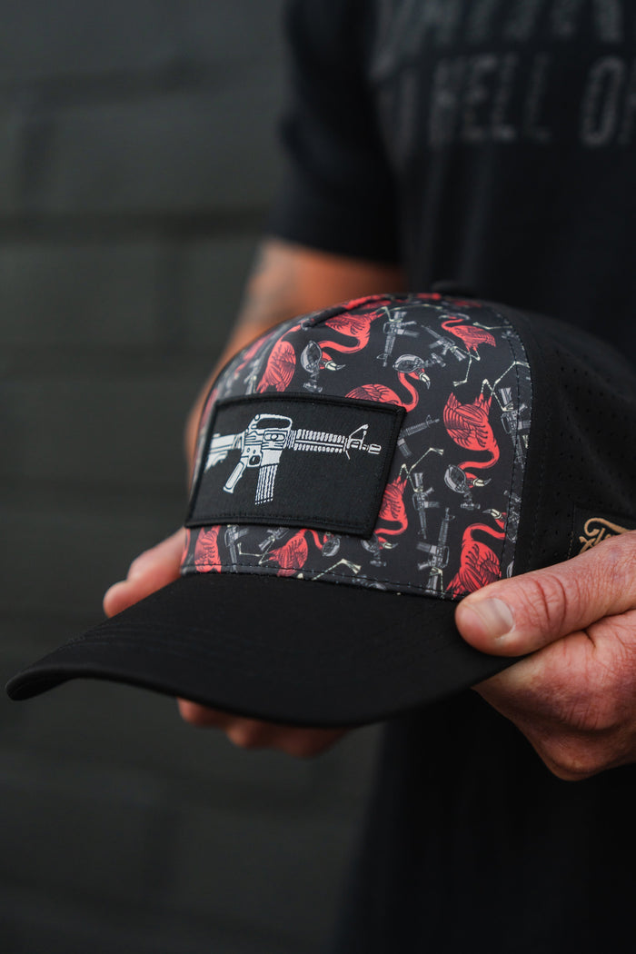 A person holds the Tactimingo Hydro Snapback Hat by Tango Charlie Apparel, featuring a black design with red flamingos and guns. It has a rectangular patch displaying a rifle and is sweat-resistant, ideal for adventures. The blurred background reveals an indistinct wall, enhancing its rugged appeal.