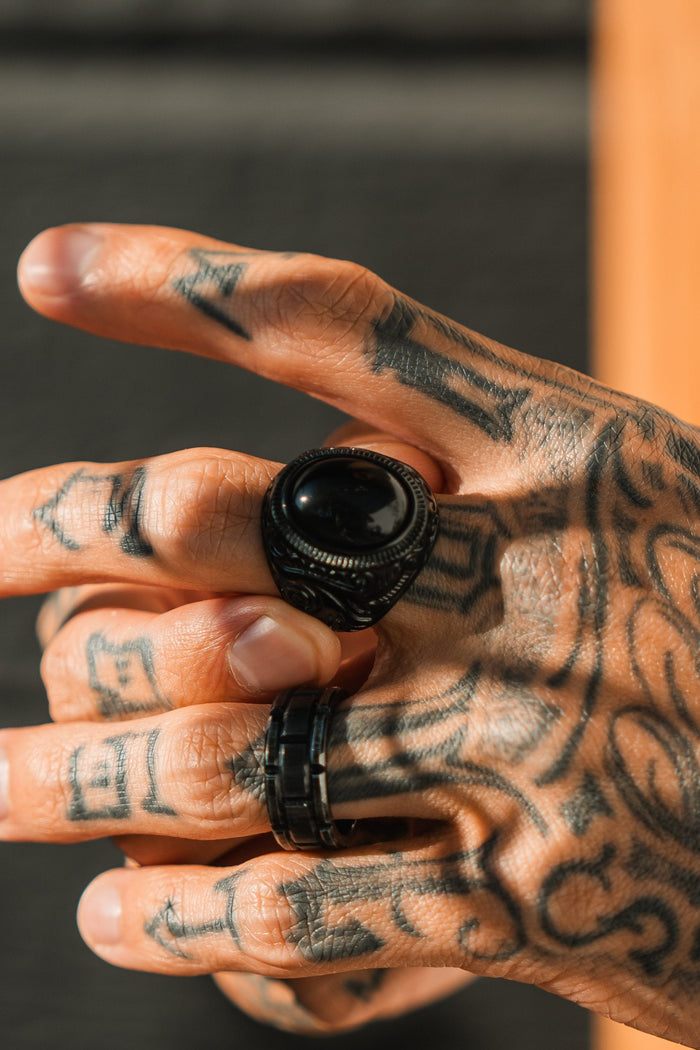 A tattooed hand is adorned with two black rings, one featuring a large round stone similar to Relentless Betrayals The Black Pearl Ring. Intricate tattoos cover the fingers and hand, casting shadows in the sunlight against a softly blurred background.