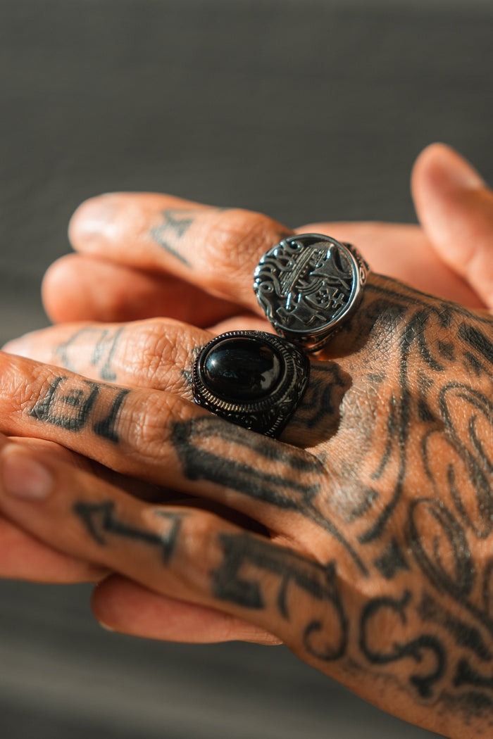 A close-up shows tattooed hands with two ornate rings: a detailed silver design and Relentless Betrayals The Black Pearl Ring, featuring a black stone in a textured band. The intricate tattoos alongside these elegant rings form a captivating tableau.