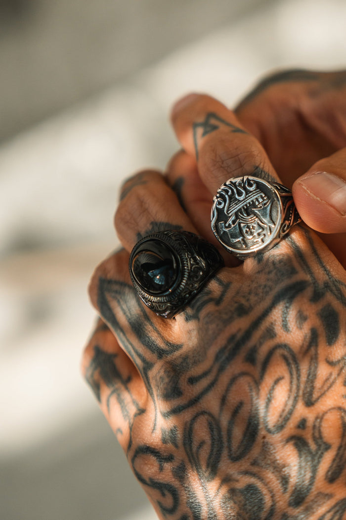 A close-up shows a tattooed hand wearing two ornate rings. One is Relentless Betrayals The Black Pearl Ring with a dark stone, and the other features an engraved face. Soft lighting highlights the detailed tattoos and intricate ring carvings.