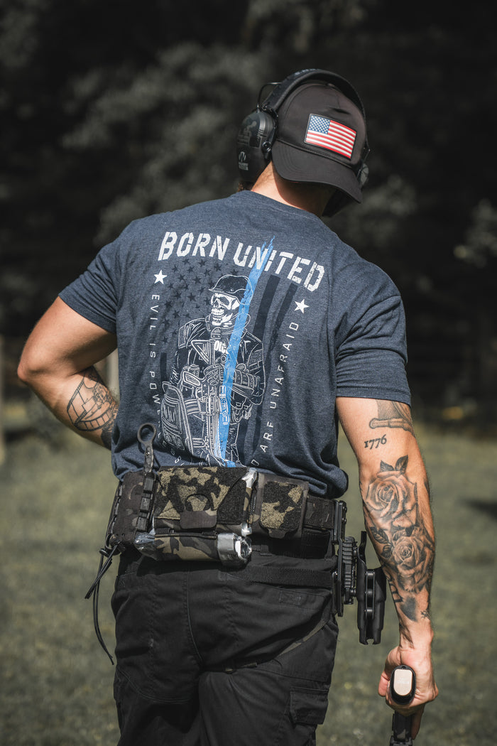 A man in The Good Are Unafraid tee by Born United Private Label, tactical gear, and an American flag cap stands behind the blue outdoors with a firearm. His tattooed arms and hearing protection show his support for those who stand behind the blue.
