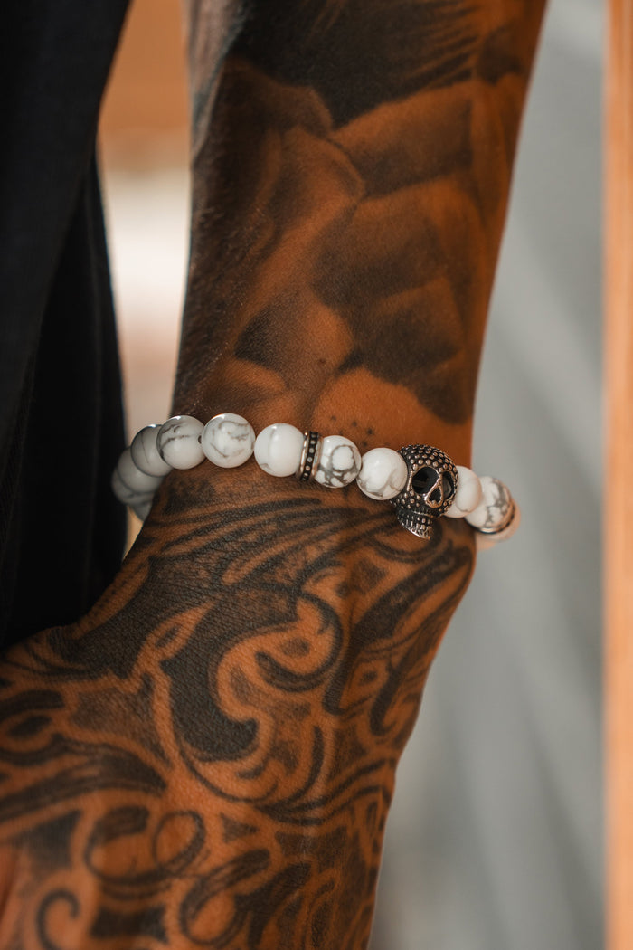 A close-up of a tattooed forearm wearing the Relentless Betrayal Until Death Storm Cloud Bead Bracelet, featuring white beads and a metallic skull charm. The tattoo displays dark, intricate patterns over most of the skin.