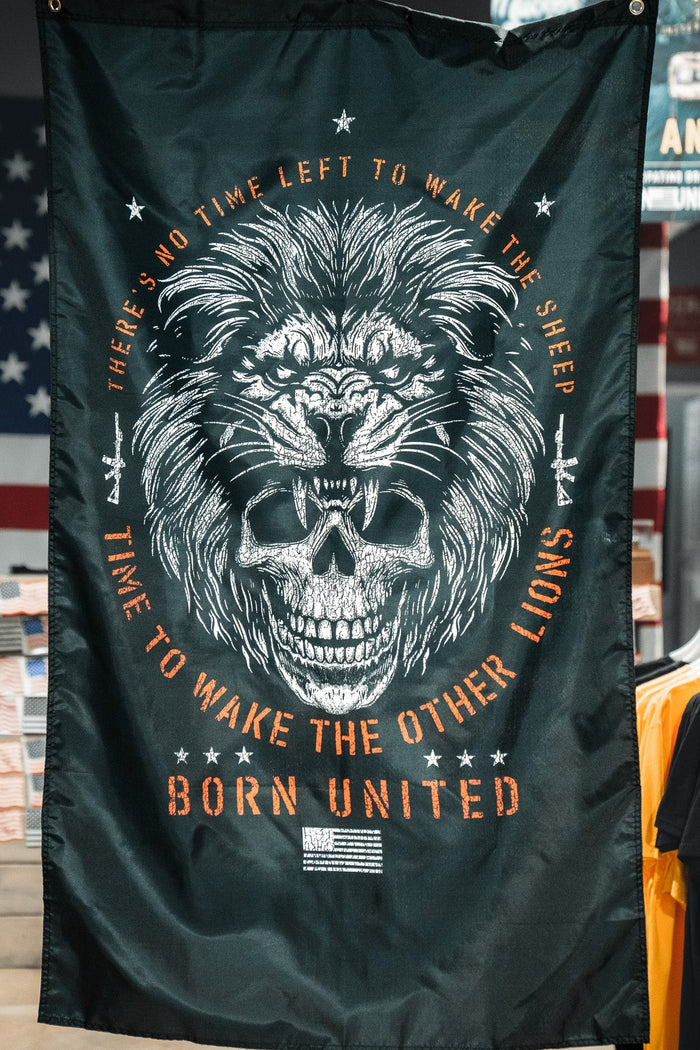 The Waking Lions Flag by Born United Private Label, crafted from durable polyester, showcases a roaring lion over a skull with the text: “There’s no time left to wake the sheep. Time to wake the other lions. Born United.” Displayed in a store setting.