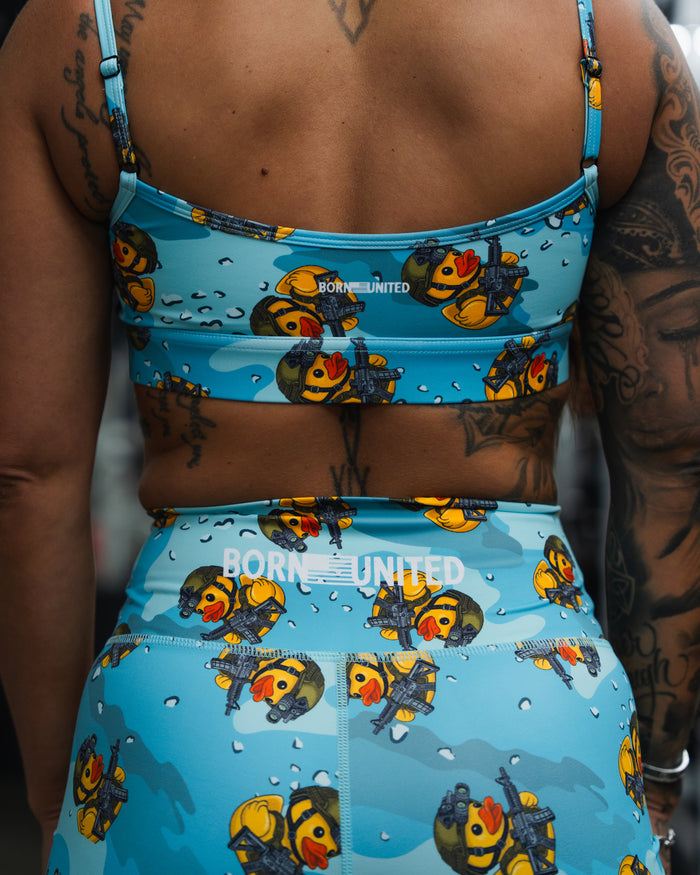 A person wears Born United's blue sports bra and leggings with a rubber duck print and "BORN UNITED" on the back, paired with Women's High-Waisted Seamless Front Compression Training Shorts by TactiQuack, showing off their tattooed arm and back.