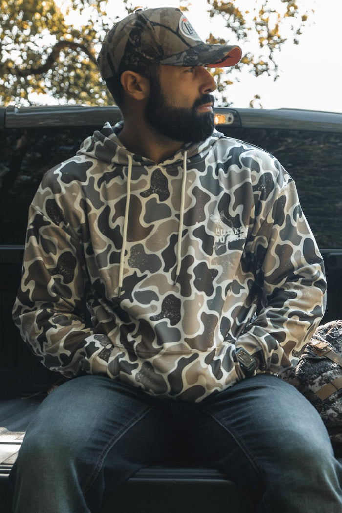 A bearded man in a Mucker Bucket Wood Ducks Premium Athletic Duck Camo Hoodie and cap sits on a pickup tailgate in sunlight, with a camo-patterned bag by his side and trees in the background.