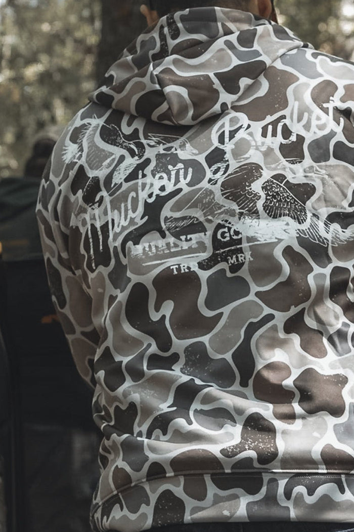 A person wearing a gray and white Mucker Bucket Wood Ducks Premium Athletic Duck Camo Hoodie with "Huckin Bucket" on the back sits outdoors in a wooded area, while another person is seated in the background.