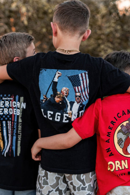 Three boys stand with their arms around each other. The central boy wears a Born United Private Label Youth Forever Legend tee featuring people, an American flag, and the word LEGEND. The boy on the right sports a red T-shirt with a circular graphic.